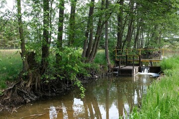 Small river flowing through wooded area in countryside. There is small bridge and culvert over water.