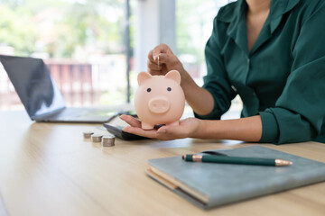 Fototapeta premium A woman is planning to save money. She puts coins in a piggy bank and records her savings in her notebook. The concept of saving money and being financially responsible. 