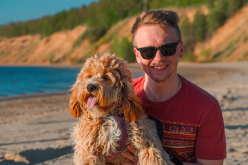 A curly-haired brown Labradoodle or Cavapoo dog with a young man are best friends by the sea at sunset.