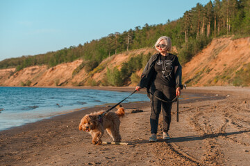 A curly-haired brown Labradoodle or Cavapoo dog with a adult woman are best friends by the sea at sunset.