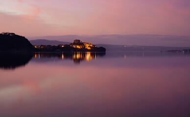 Naklejka premium Sunset over Lake Bolsena paints the sky with warm hues, reflecting on tranquil waters. A serene Italian landscape perfect for travel, nature, and romantic visual themes. Peaceful and captivating views