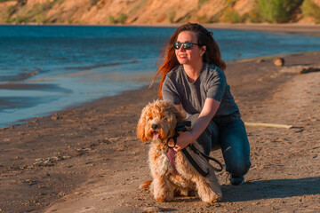 A curly-haired brown Labradoodle or Cavapoo dog with a young woman are best friends by the sea at sunset.