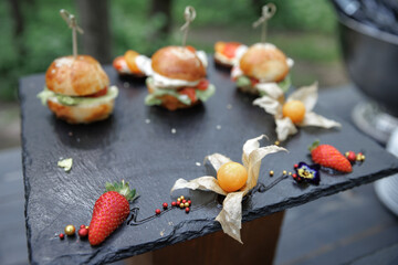 Elegant presentation of gourmet mini burgers on a slate tray, garnished with fresh strawberries and physalis. Ideal for upscale events, showcasing culinary artistry and premium ingredients.