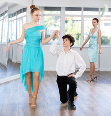 Naklejka premium Adolescent dancers rehearsing ballroom dance as couple under guidance of female coach in choreography studio. Elegant girl in blue dress gracefully stepping around boy standing on knee on dance floor