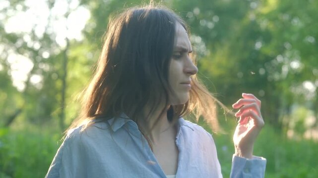 Young woman experiencing intense mosquito bite discomfort, swatting and scratching while surrounded lush park greenery during sunny summer day. Woman feeling discomfort from mosquitoes bites in nature