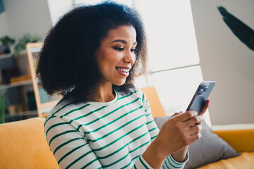 Young woman in a cozy living room enjoying leisure time, smiling while using her smartphone,...