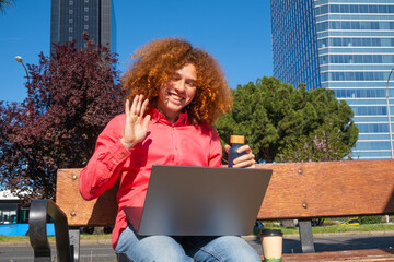 Young man with curly hair waving during a video call, working outdoors with laptop and reusable cup. Concept of remote work, green lifestyle and sustainability