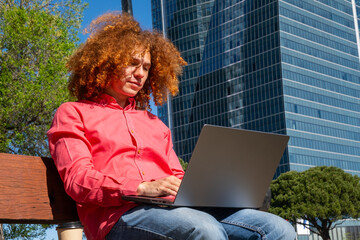 Low angle portrait of a Young man with curly red afro hair working on a laptop in the city, sitting on a bench. Remote work and digital nomad lifestyle concept. Office building on the background