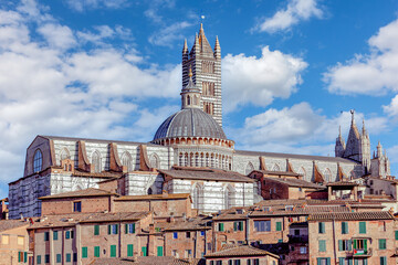 The grand Siena Cathedral towers over the medieval buildings of Siena, showcasing its stunning architecture against a backdrop of fluffy clouds and a bright blue sky.