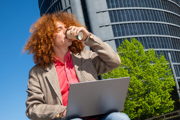 Young man with curly hair drinking from a reusable cup while typing on a laptop outdoors on a financial district. Concept of sustainable remote work, eco habits and modern business lifestyle