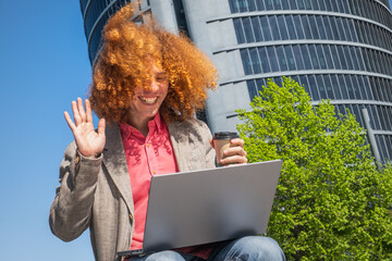 Young man with curly hair waving during a video call, working outdoors with laptop and reusable cup. Modern office building in the background. Concept of remote work and sustainable lifestyle