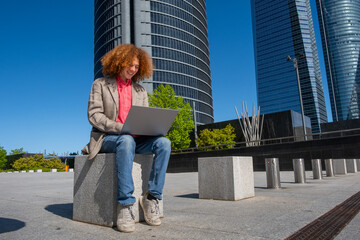 Smiling young man with curly red hair working outdoors on a laptop in front of corporate skyscrapers. Dressed in a casual blazer, he represents modern freelance work and eco-conscious business living