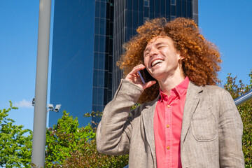Smiling young man with curly hair having a phone conversation in a financial district, dressed in a blazer. Modern glass towers behind him. Concept of freelance work and urban life