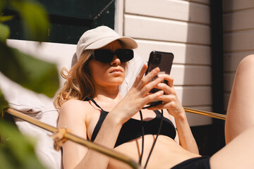 Calm cool blonde woman sitting and tanning on backdoor house, wear sunglasses, cap and swimsuit. Woman messaging, typing sms, read news, working online using mobile phone.
