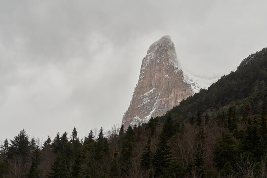 A solitary, sharp rock formation towers above dense pine forest in the Ordesa Valley, Aragonese Pyrenees. Snow covers the mountain slope under a moody winter sky with clouds surrounding the summit.