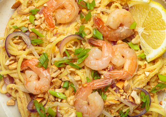 Close up of delicious pad thai noodles garnished with cooked shrimps, roasted peanuts, chopped green onions, red onion rings and a lemon wedge, traditional dish of Thailand