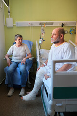 Senior man with broken leg or knee replacement surgery sitting on hospital bed talking to his wife