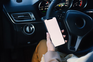 A woman in the driver's seat of a car holding a mockup of a smartphone with a white blank screen. The concept of mobile communication, driving with technology and navigation on the road.