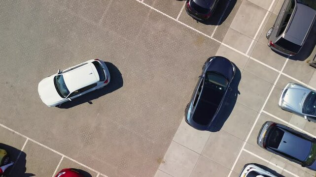 Aerial footage of a parking lot with several cars parked in designated spaces, including a white car maneuvering. The lot has both paved and concrete sections under clear sunlight.
