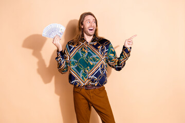 Happy man in stylish outfit holding cash and pointing to the side with excitement on a beige...