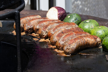 Several sausages side by side roasting on top of a large griddle outdoors