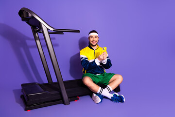 Smiling young man in a retro-style outfit sitting next to a treadmill on a violet background holding a phone