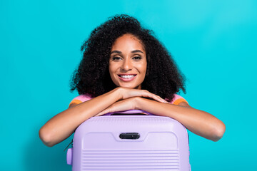 Smiling young woman leaning on a purple suitcase against a teal background promoting travel and...