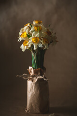Wilting daffodils with yellow and white petals and green stems bundled in brown paper tied with twine, creating a melancholic still life against a brown backdrop