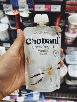 Vanilla-flavored Chobani Greek Yogurt in a squeezable pouch, held in hand in front of a refrigerated shelf. White packaging with vanilla flowers. Concept: dairy snack, portable yogurt.