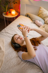 A teenage girl with headphones on her head uses a mobile phone in the bedroom. She's lying on the bed.