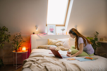 A young woman uses a laptop and writes in a notebook while sitting on her bed at home. Headphones on her head.