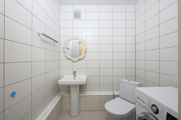 bright bathroom features white tile walls, sink, toilet, and washing machine. A decorative mirror...