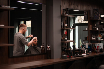Barber trims a seated male client’s hair while reflected in a large mirror, surrounded by shelving with grooming tools and modern barbershop décor..