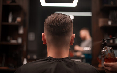 Barber sprays styling product onto the back of a male client’s fresh fade haircut in a modern...