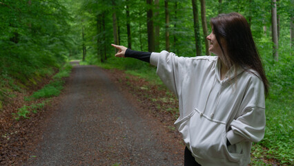 A young woman in a light beige hoodie in the forest pointing toward the road.