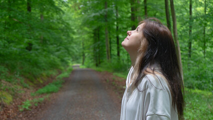 A young woman in a light beige hoodie in the forest looking up at the sky with a forest path in the...