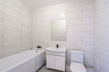 Modern bathroom with white marble tiles, white bathtub, sink with cabinet, and toilet. Clean and...