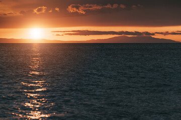 Golden sunset reflecting on calm sea with distant mountains and clouds creating a dramatic and peaceful atmosphere