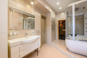 Bright bathroom with beige tiles, white vanity, large mirror, shower cabin, and sauna entrance