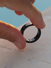 Hand holds black smart ring near pool with health tracker.