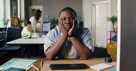 An employee looks at the camera sitting at his desk in the office. His colleagues are moving around...