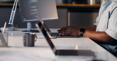 A thoughtful programmer uses a laptop and a computer. He's sitting at his desk in the office.