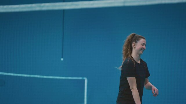Portrait: red-haired smiling girl playing badminton indoors. Happy female athlete hits shuttlecock with a racket across a net. Practicing serve before playing in badminton on inside court