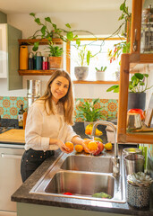 Smiling Light-Skinned European Woman Washing Citrus Fruits in Bright Home Kitchen with Houseplants and Natural Light