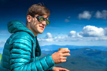 Outdoor adventure man in sunglasses and turquoise jacket drinking coffee and smiling with mountain...