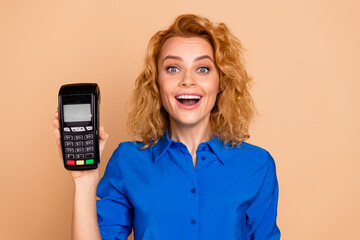 Smiling professional woman displaying a payment terminal in a blue shirt against a beige background
