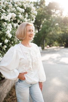 Stylish mature woman 55-59 year old wearing white top over rose flowers with city view outdoors.