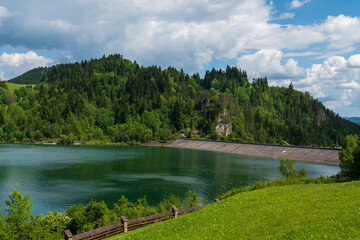 Wide view of Czorsztyn Lake dam in Poland with surrounding green forest and hills. Concrete slope structure meets calm water under partly cloudy sky.
