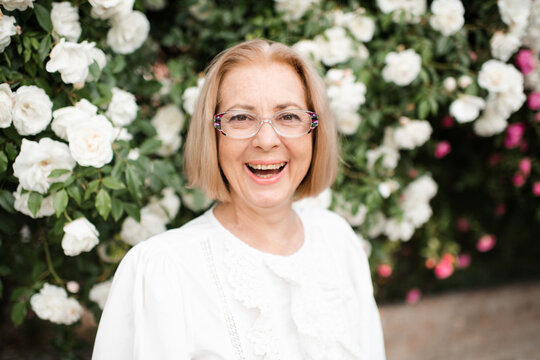 Happy blonde mature woman 55-59 year old wearing eye glasses over blooming bushes with rose flowers outdoors. Summer season. Happiness.