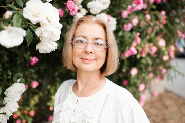 Smiling blonde mature woman 55-59 year old wearing eye glasses over blooming bushes with rose flowers outdoors. Healthy lifestyle.